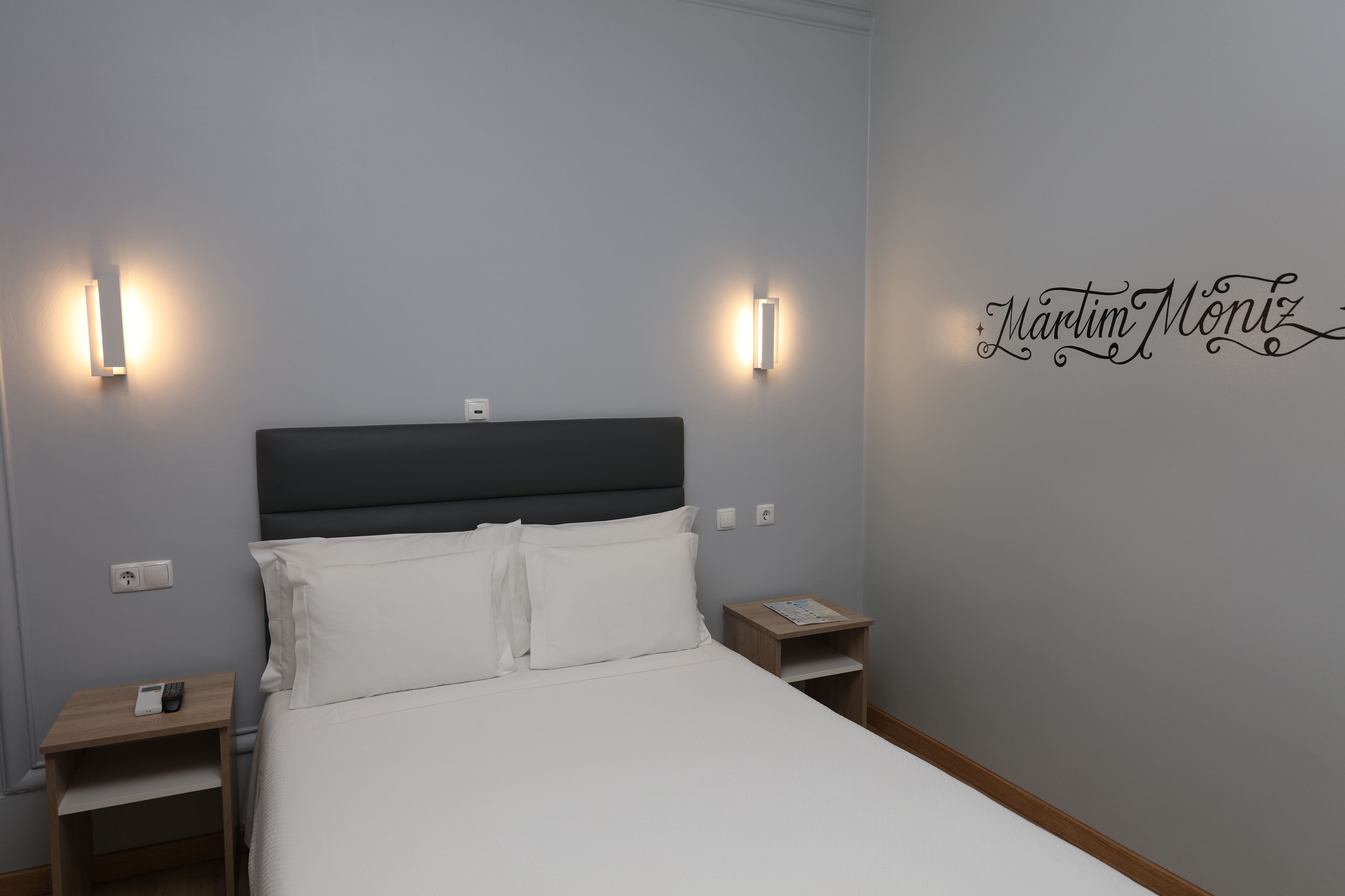 A neatly made bed with white linens and two bedside tables, one with a phone and the other with a lamp.