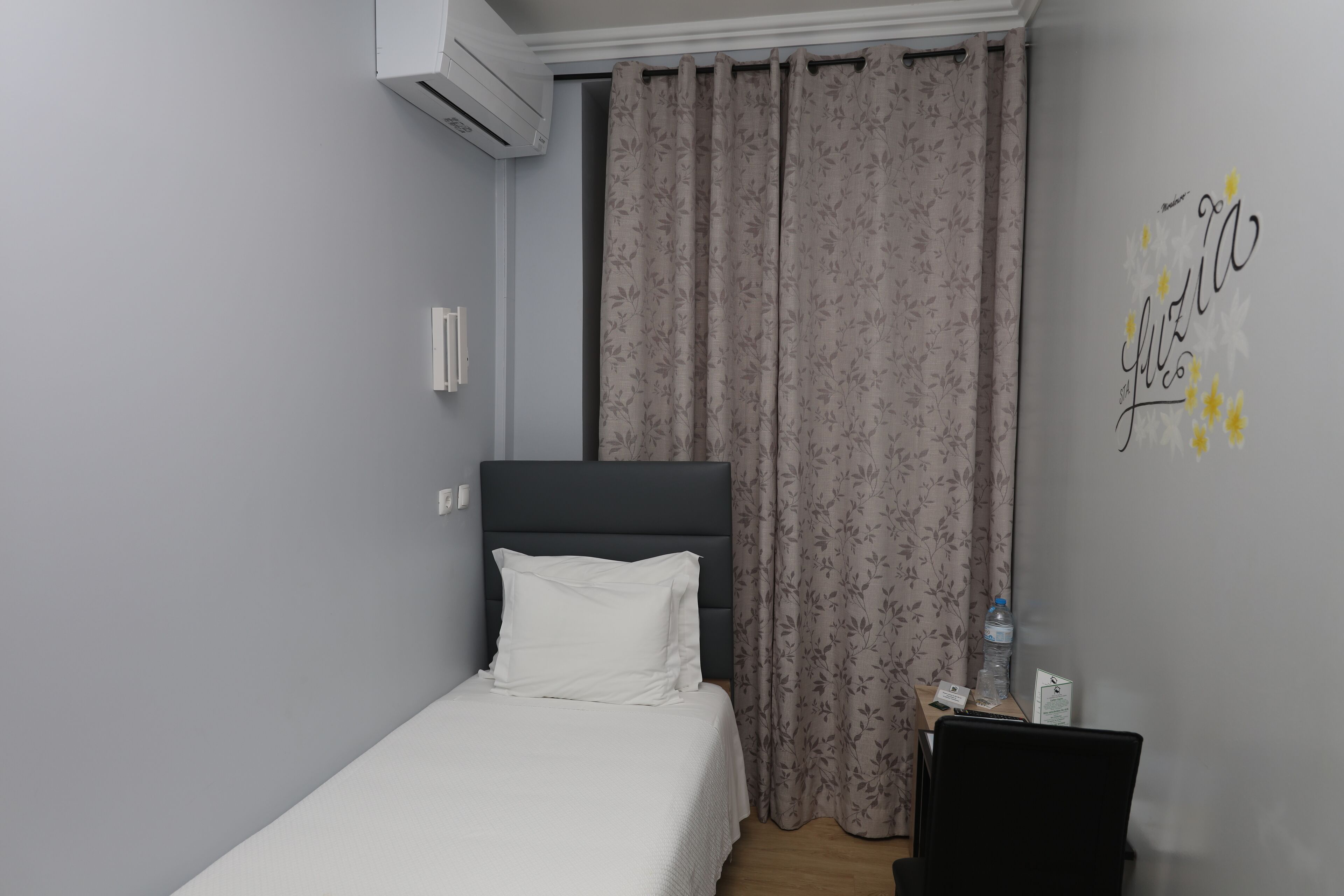 Une chambre d’hôtel comprenant un lit, un bureau, une chaise, une bouteille d’eau et un mur décoré avec un nom.