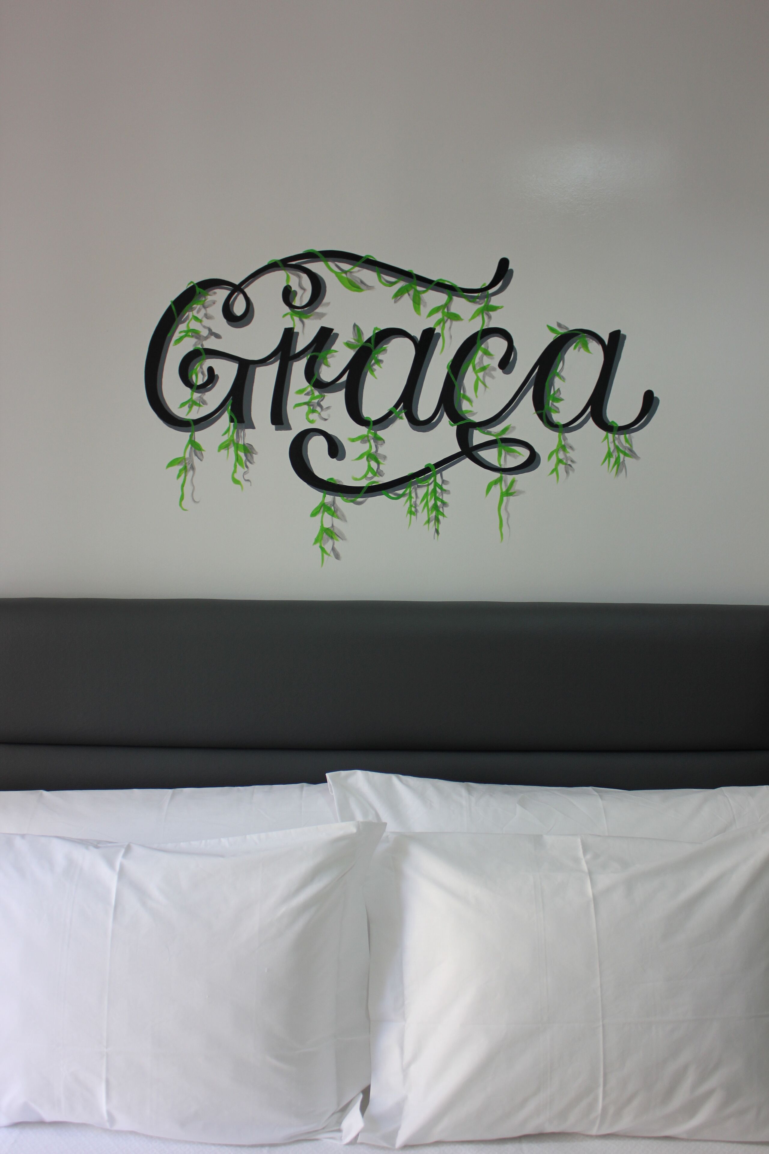 A wall with the name "Graca" in decorative script, above a bed with white bedding.