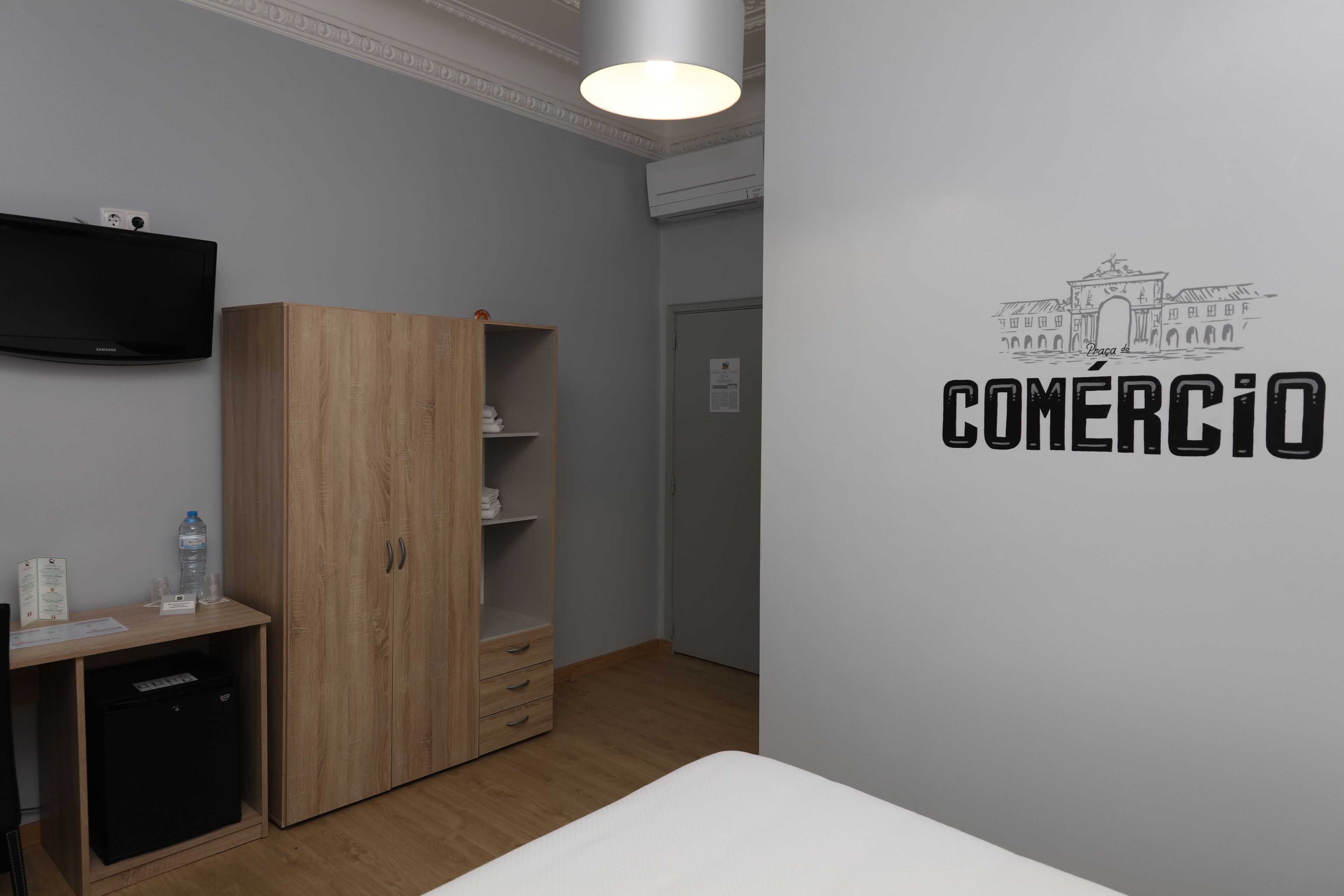 A hotel room with a wooden wardrobe, a desk with a water bottle, a TV, and a wall with the word "COMERCIO".