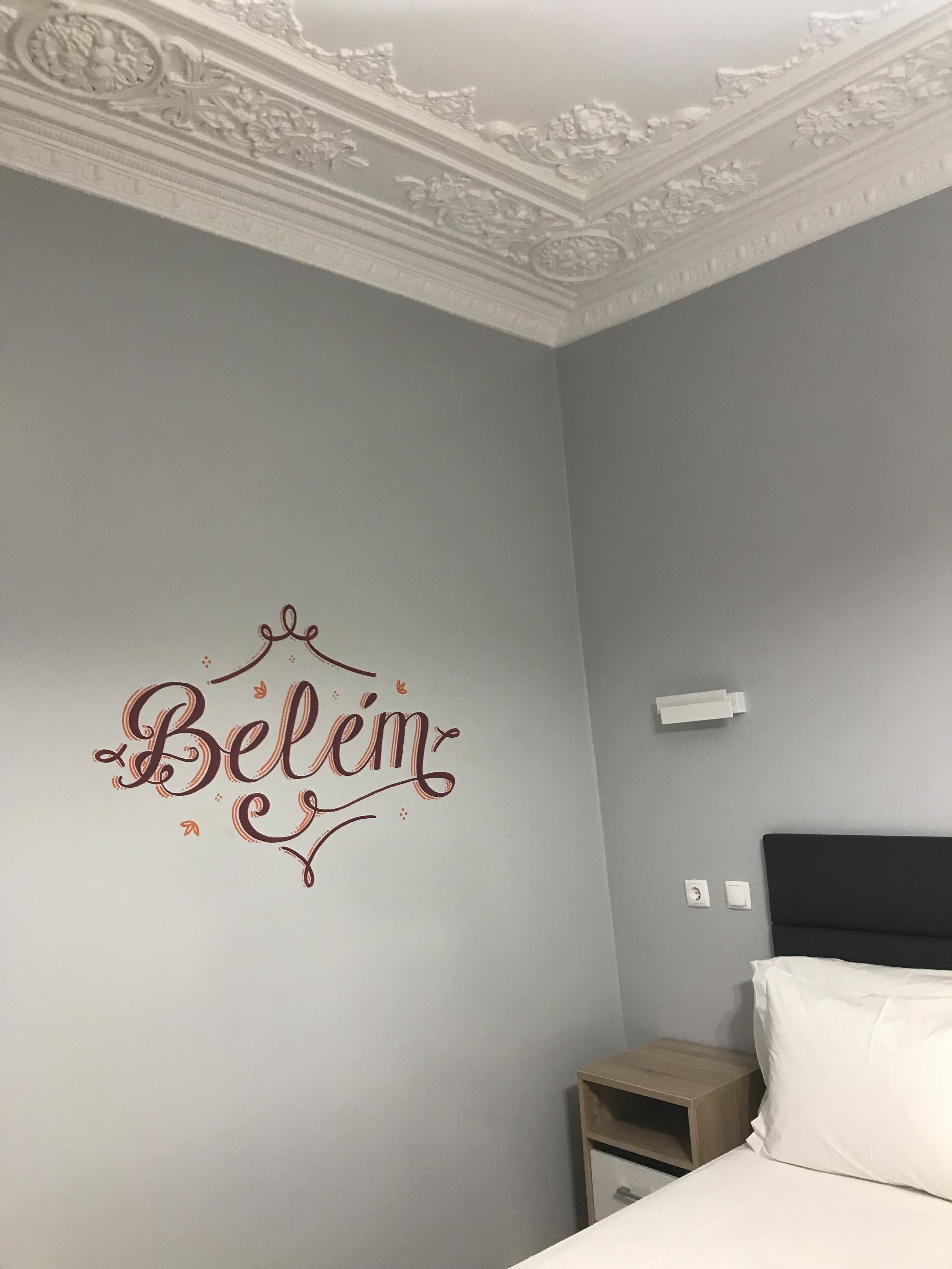 Una habitación de hotel con un techo decorativo, una cama con ropa de cama blanca, una mesita de noche y una pared con un cartel que dice 'Belém'.