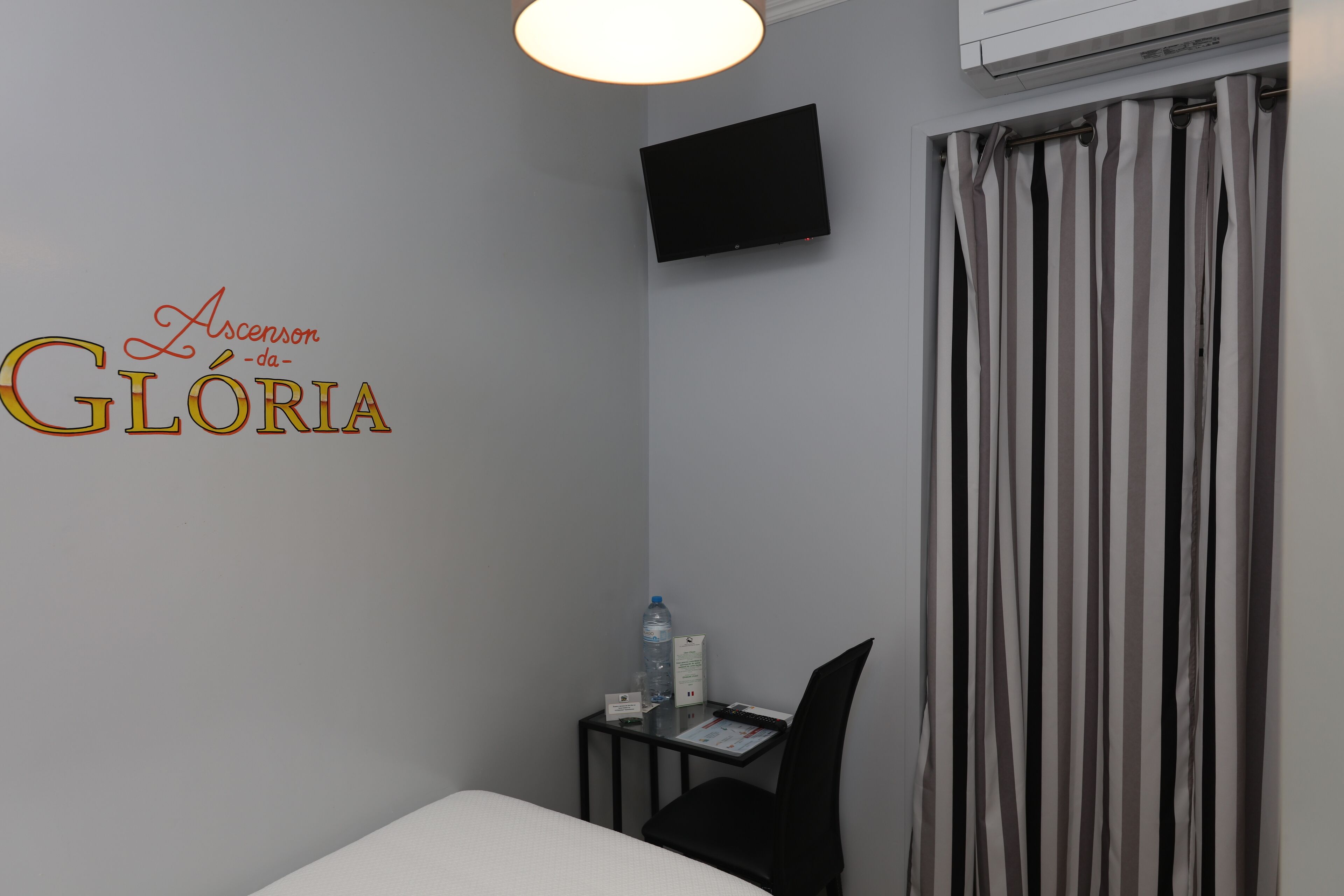 A hotel room with a bed, a desk with a chair, a water bottle, a glass of water, a flat-screen TV mounted on the wall, and a striped curtain.