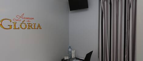 A hotel room with a bed, a desk with a chair, a water bottle, a glass of water, a flat-screen TV mounted on the wall, and a striped curtain.