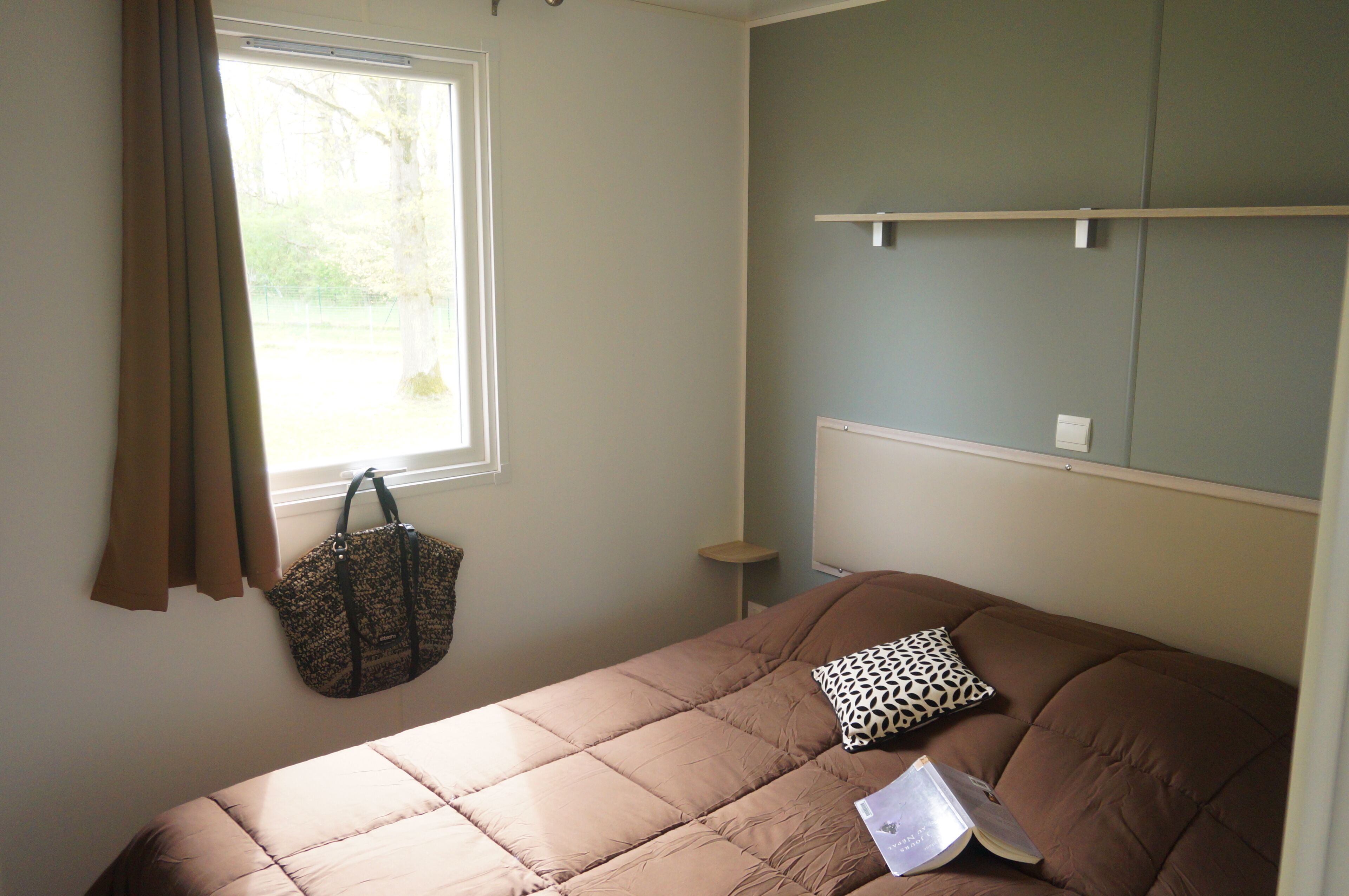A bedroom with a bed, a window with curtains, a bedside table, and a wall-mounted shelf.