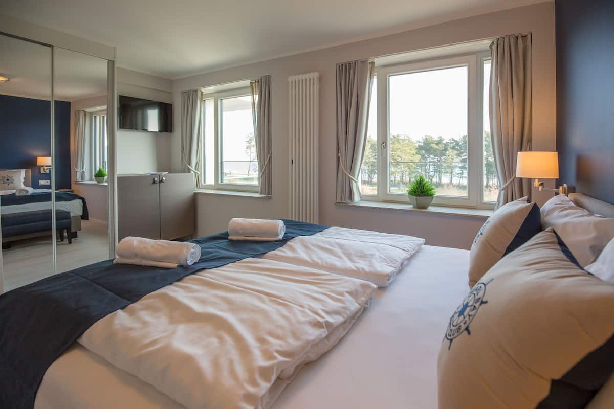 Ett hotellrum med en stor säng, en platt-TV, ett fönster med gardiner, en liten växt och ett skrivbord med en lampa.