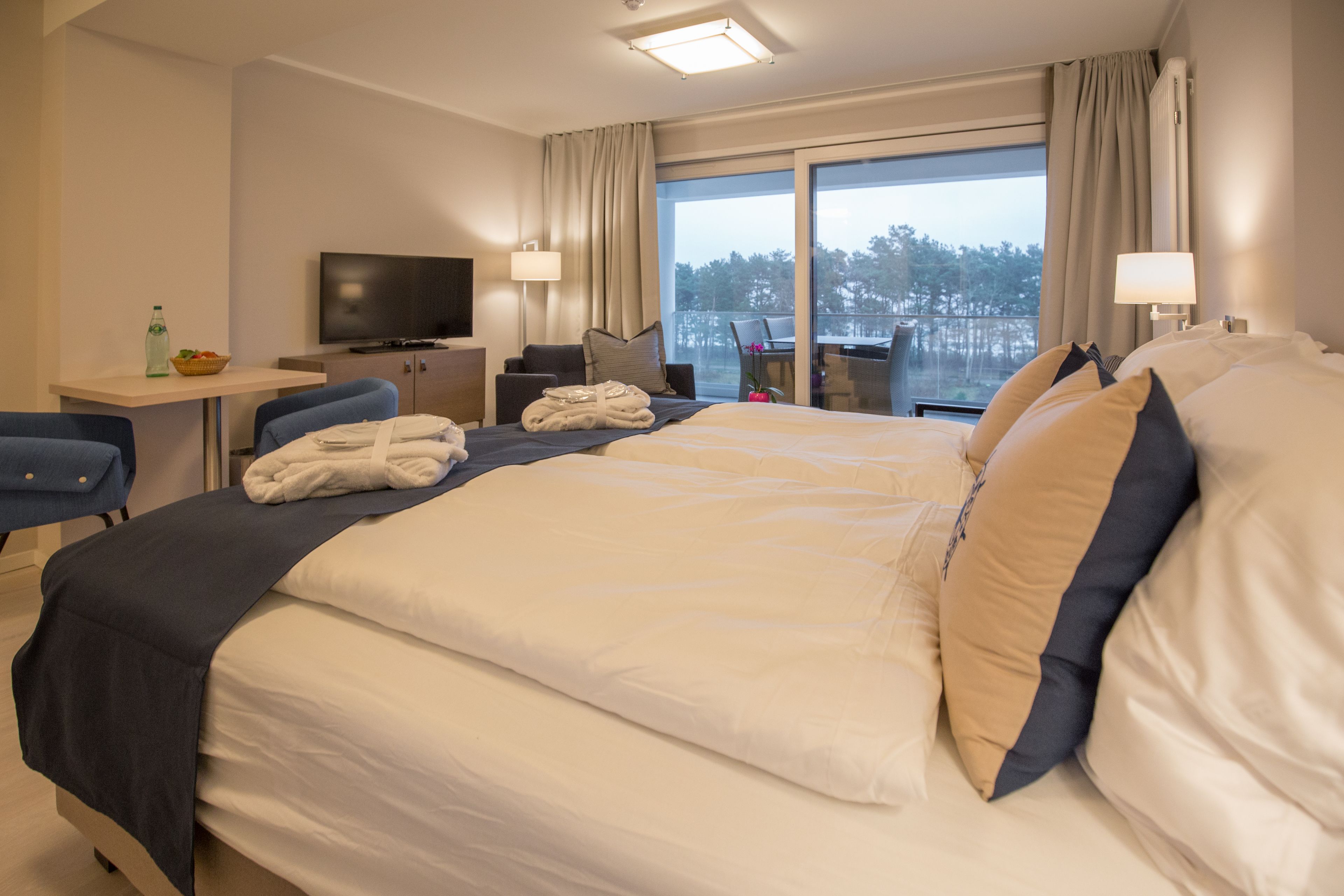 Habitación de hotel con una cama grande, un escritorio con televisor y un balcón con vista a árboles.
