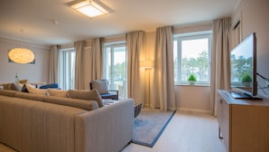 Una sala de estar moderna con un sofá, un televisor, una lámpara y vistas al exterior a través de amplios ventanales.