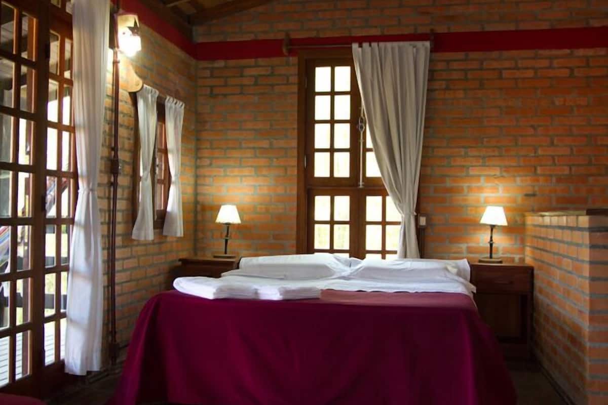 A room with a brick wall, a bed with a red bedspread, two bedside lamps, and a window with curtains.