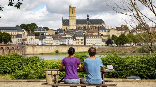 Two people sitting on a bench facing a river with a historic building in the background. - Camping de Nevers (Nevers)