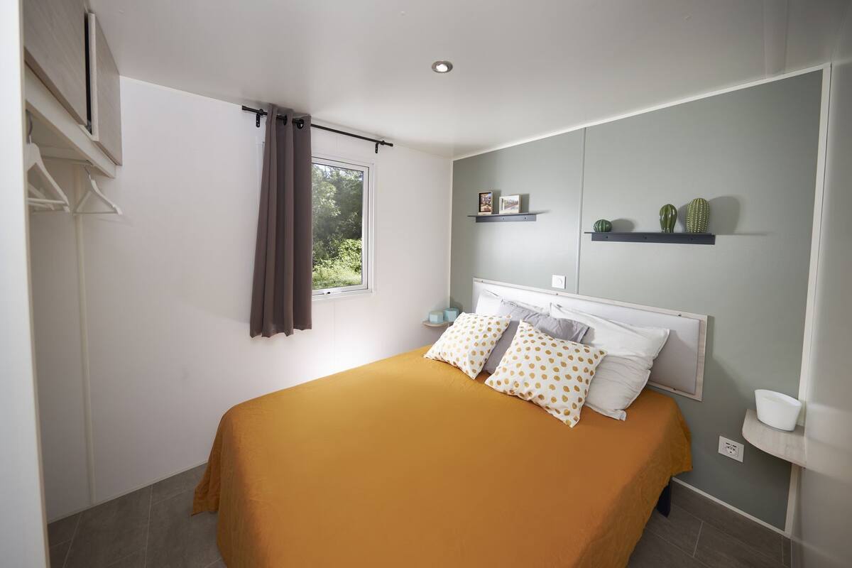 A bedroom with a bed, a window with curtains, a bedside table, and a shelf with decorative items.