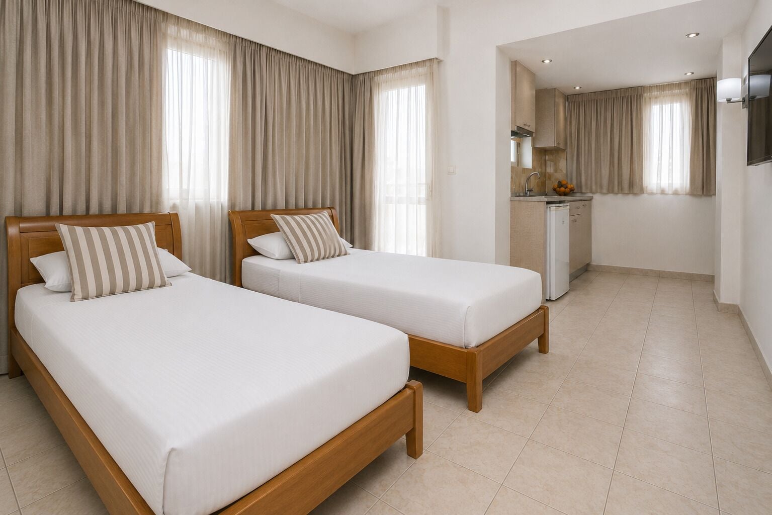 Quarto de hotel com duas camas de solteiro, cozinha compacta e televisão.