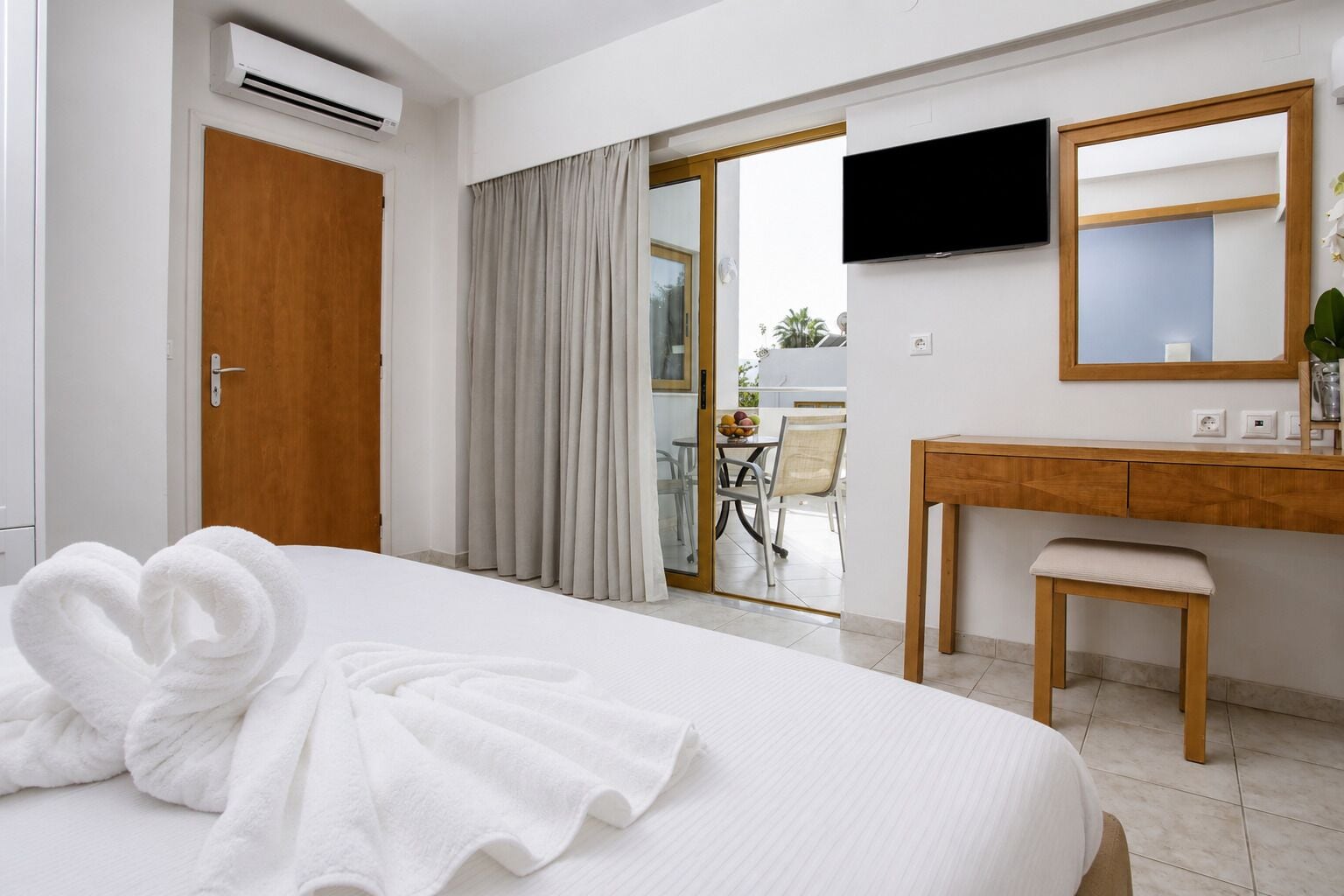 Habitación de hotel con cama, puerta de madera, unidad de aire acondicionado, baño con tocador de madera, televisión y puerta corrediza de vidrio que da a un espacio exterior.