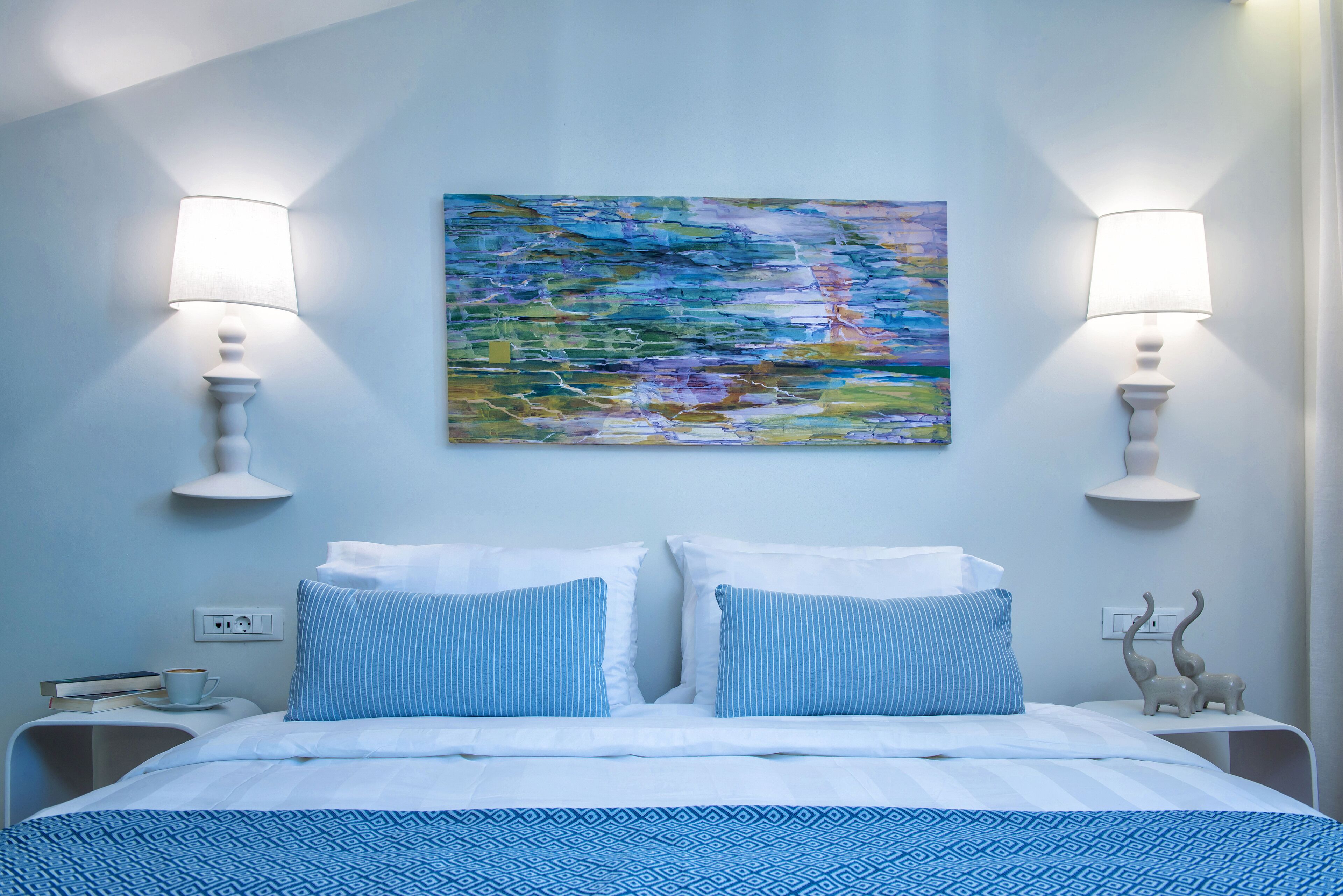 A hotel room with a bed, two bedside lamps, a painting, and a small statue on the nightstand.