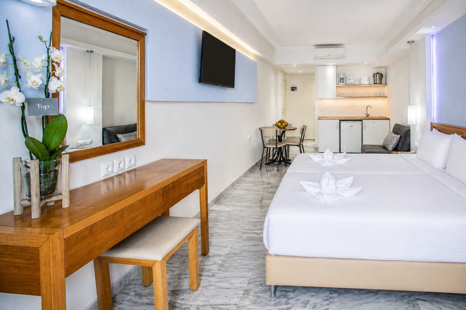 A hotel room with a wooden desk, a large bed, a mirror, a small dining table, and a kitchenette in the background.