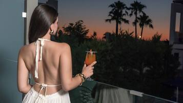 A person holding a drink on a balcony with a view of palm trees and a sunset.