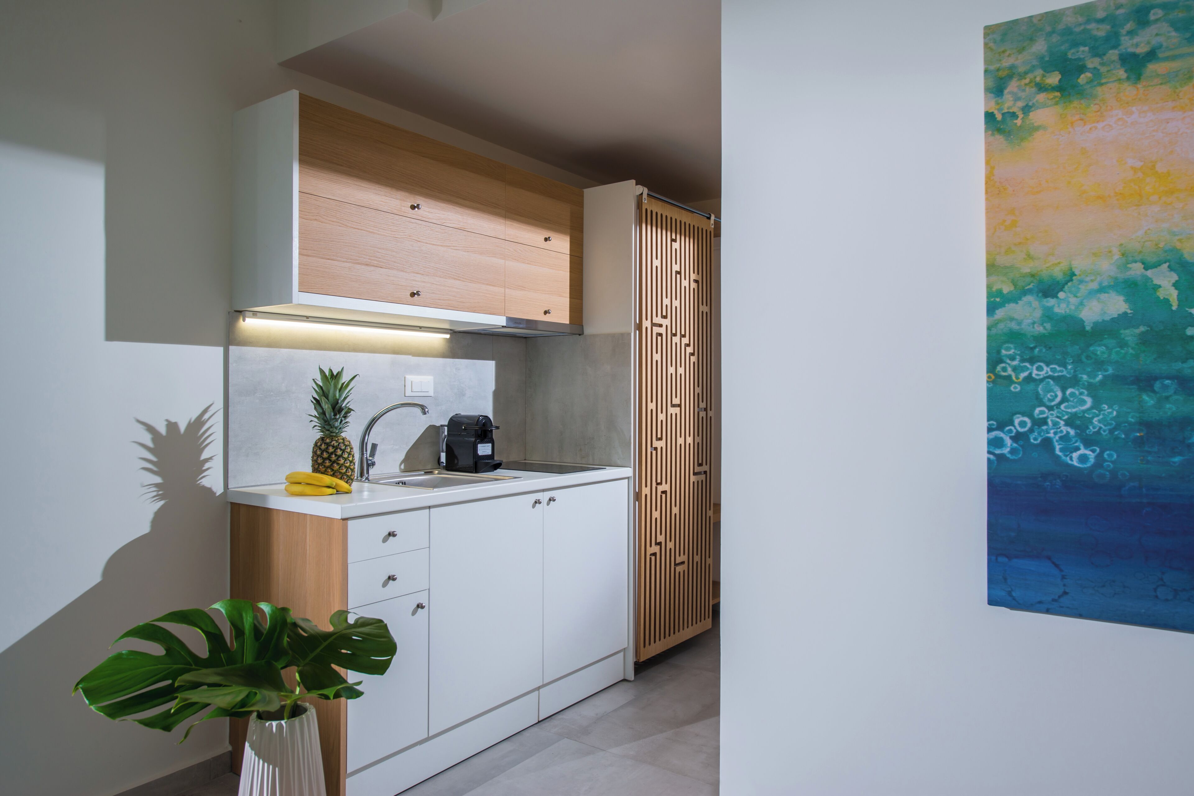 A compact kitchen with white cabinetry, a wooden upper shelf, a stainless steel sink, and a modern pendant light.