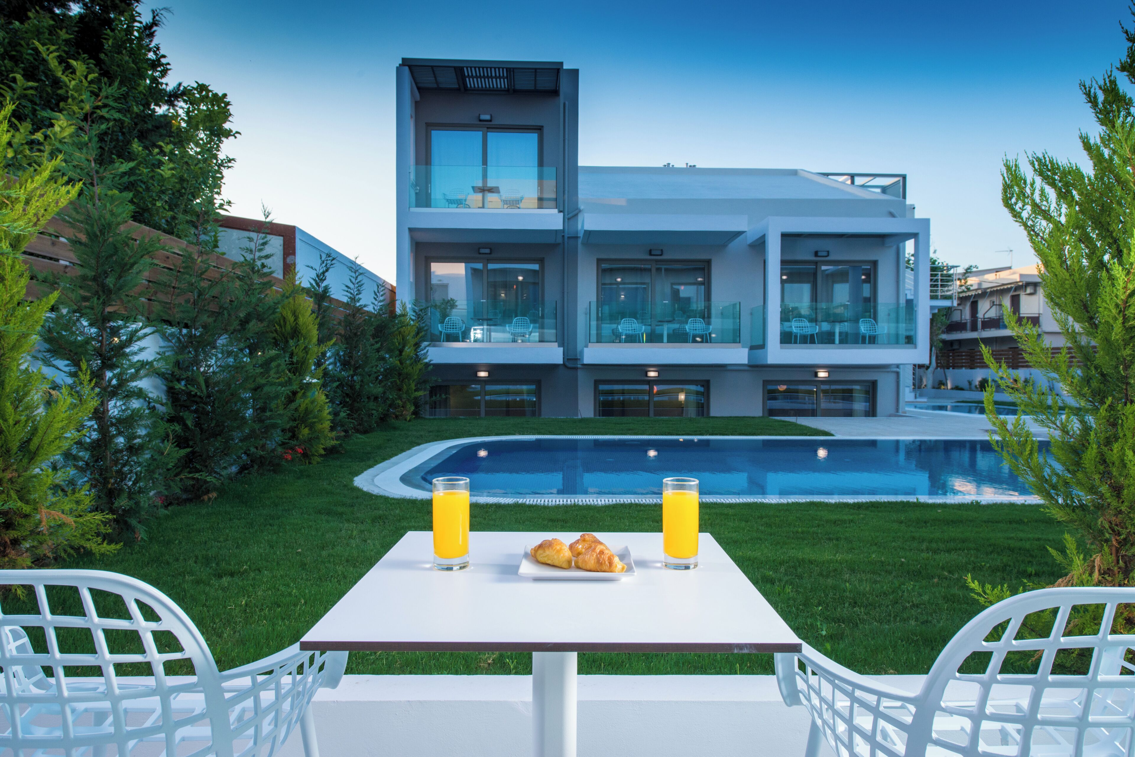 Una casa moderna amb piscina, dues taules blanques amb cadires i un croissant sobre una d'elles.