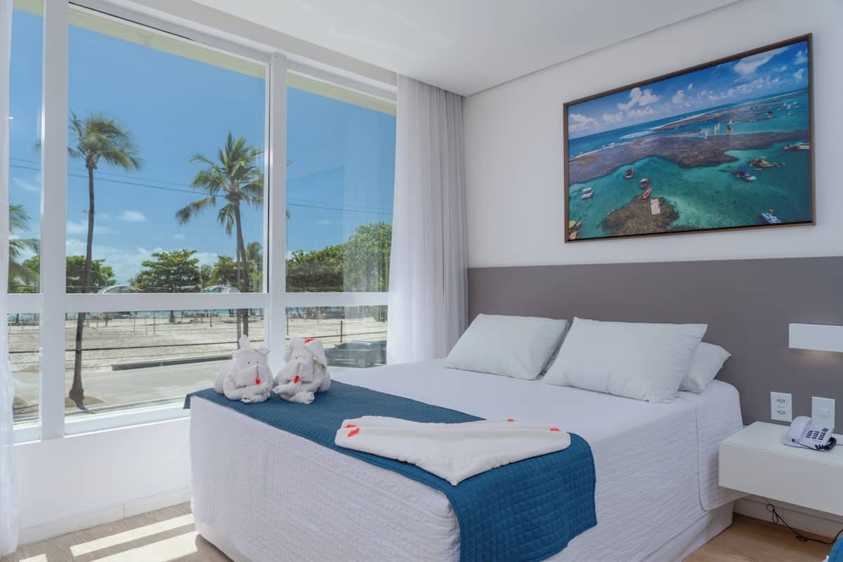 Een hotelkamer met een groot raam, een bed met wit beddengoed, een schilderij van een strand en een kleine tafel met een telefoon.