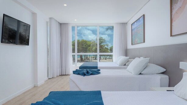 Ein Hotelzimmer mit zwei Betten, einem großen Fenster mit Blick auf einen Strand und einem Flachbildfernseher an der Wand.
