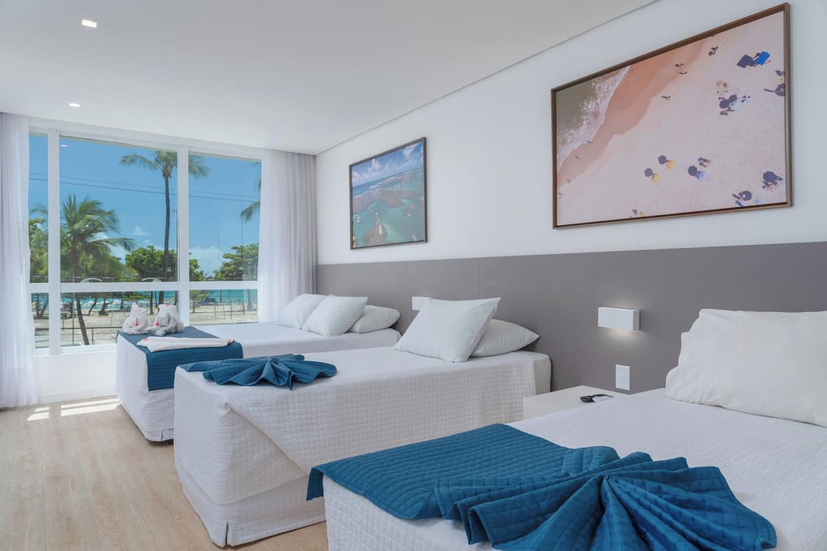 Een hotelkamer met twee bedden, een groot raam met uitzicht op palmbomen en een strand, en een schilderij van een zandstrand aan de muur.