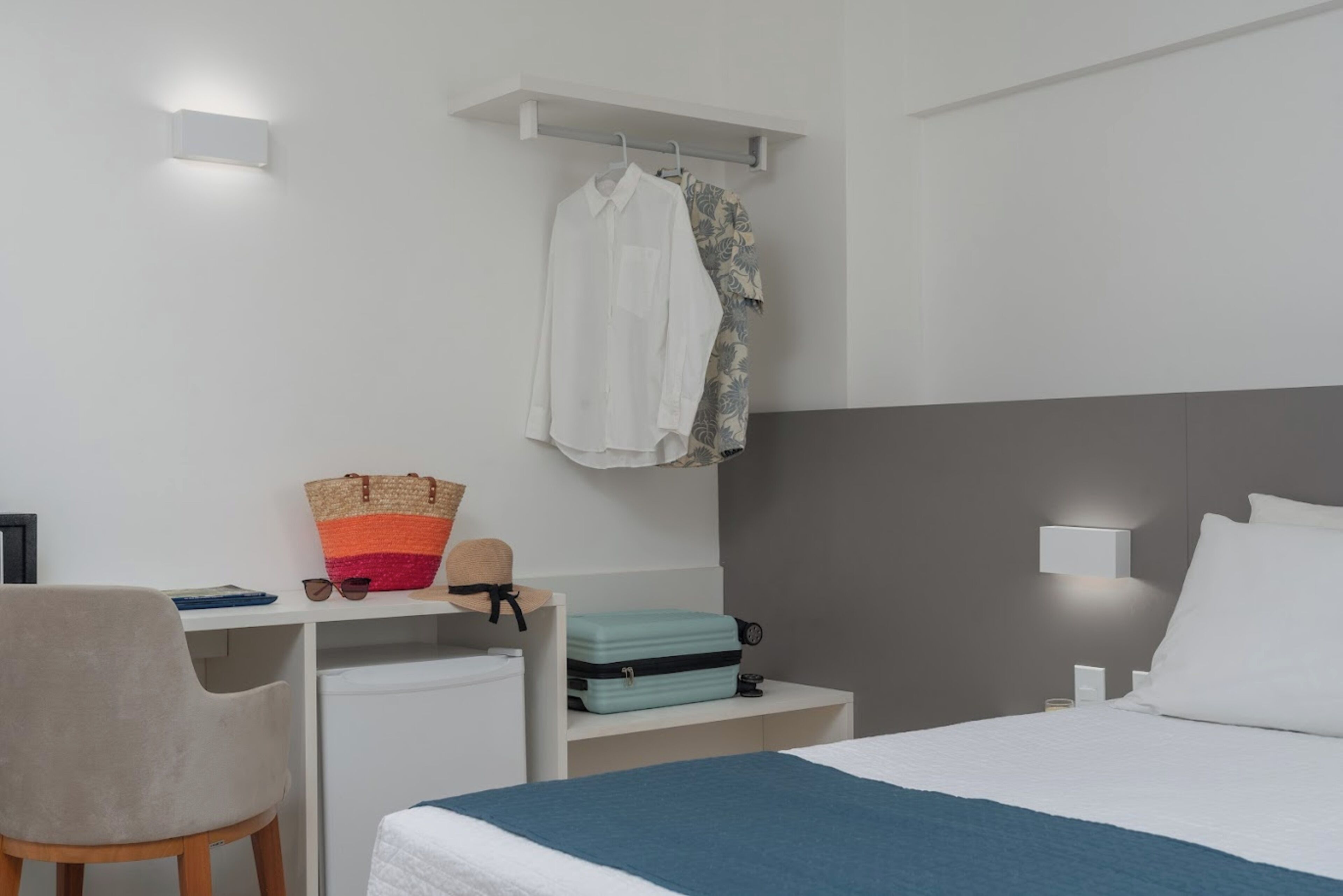 Habitación de hotel con cama, escritorio, silla, maleta y ropa colgada.
