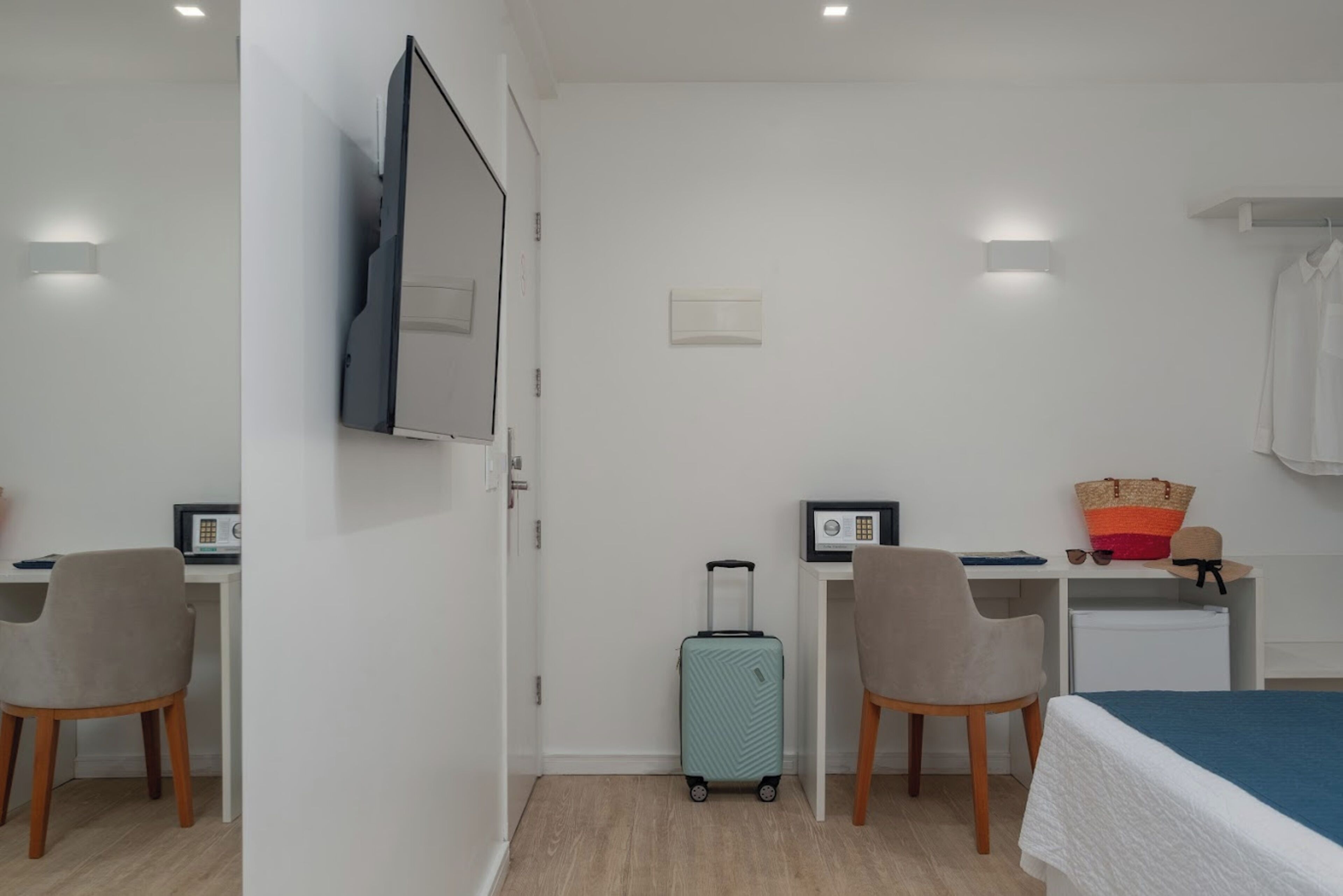 Una habitación de hotel con una cama, un escritorio con computadora, una silla, una maleta y un televisor montado en la pared.