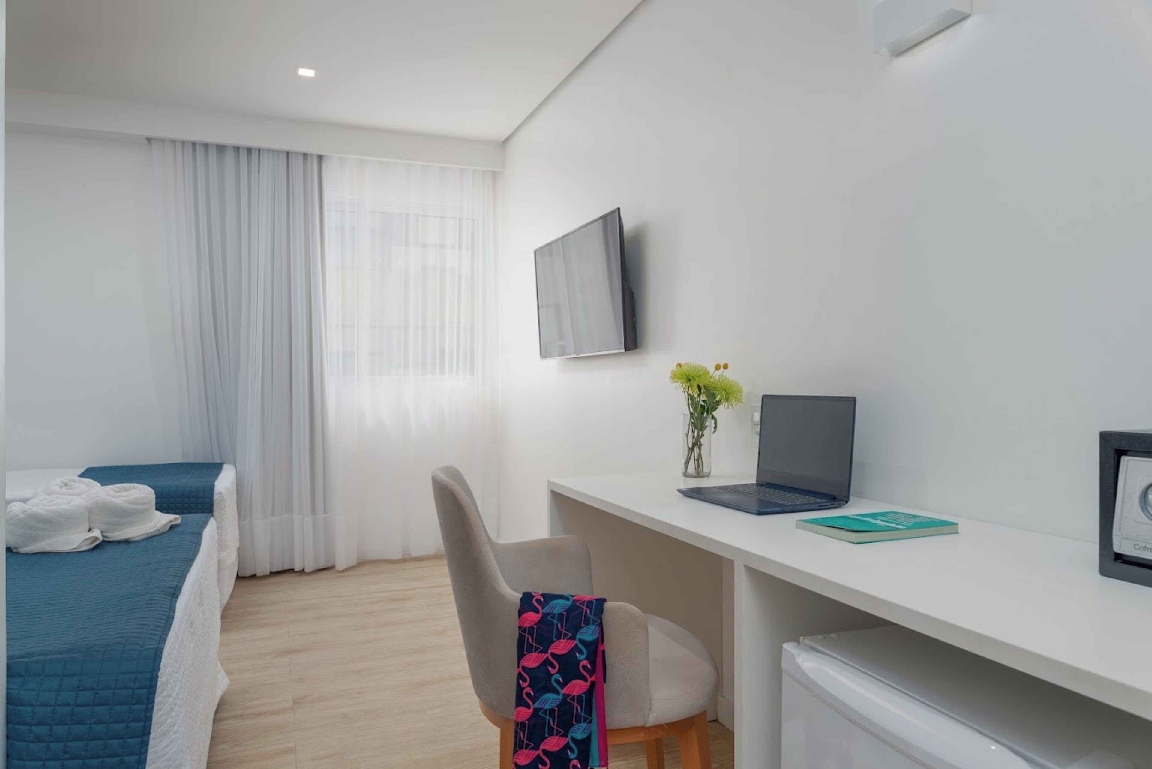A hotel room with a bed, a desk with a laptop, a chair, and a TV on the wall.