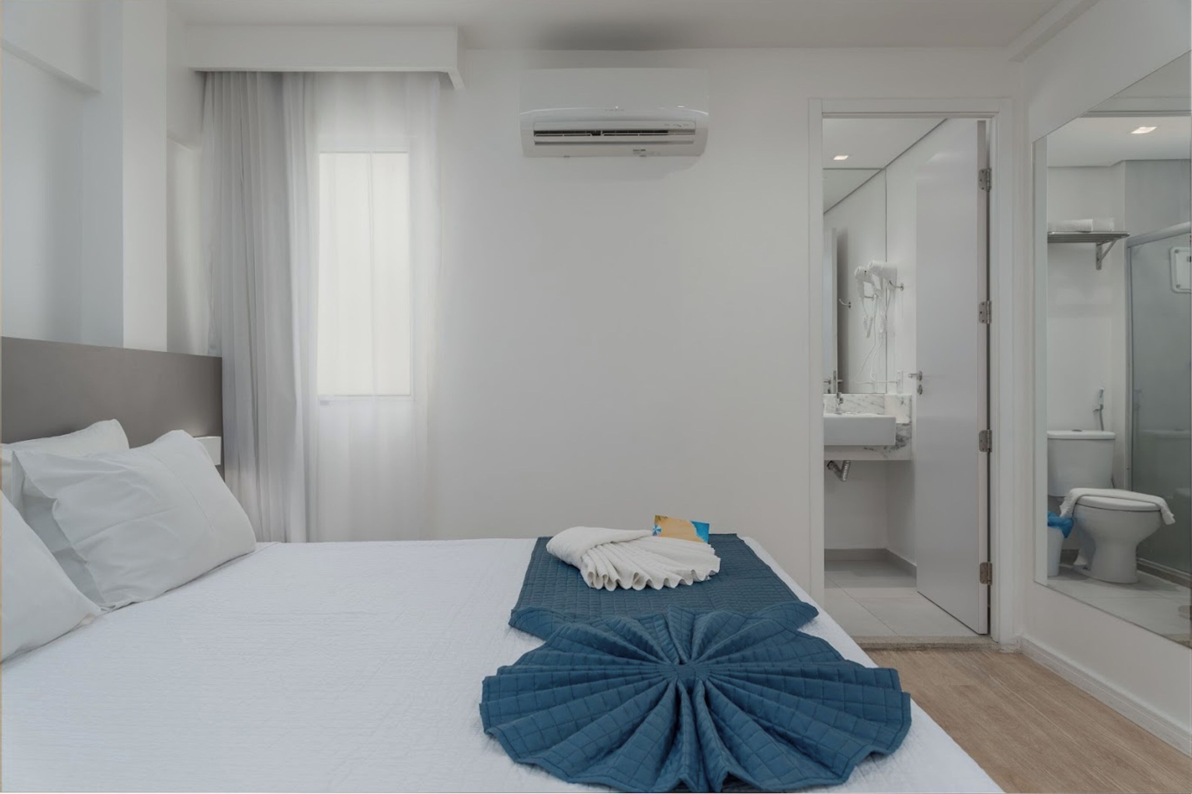 Quarto de hotel com cama, banheiro com chuveiro e vista para o banheiro através de uma porta de vidro.