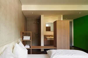 Quarto de hotel moderno com cama, prateleira de madeira com toalhas, área de chuveiro e uma parede verde decorativa.