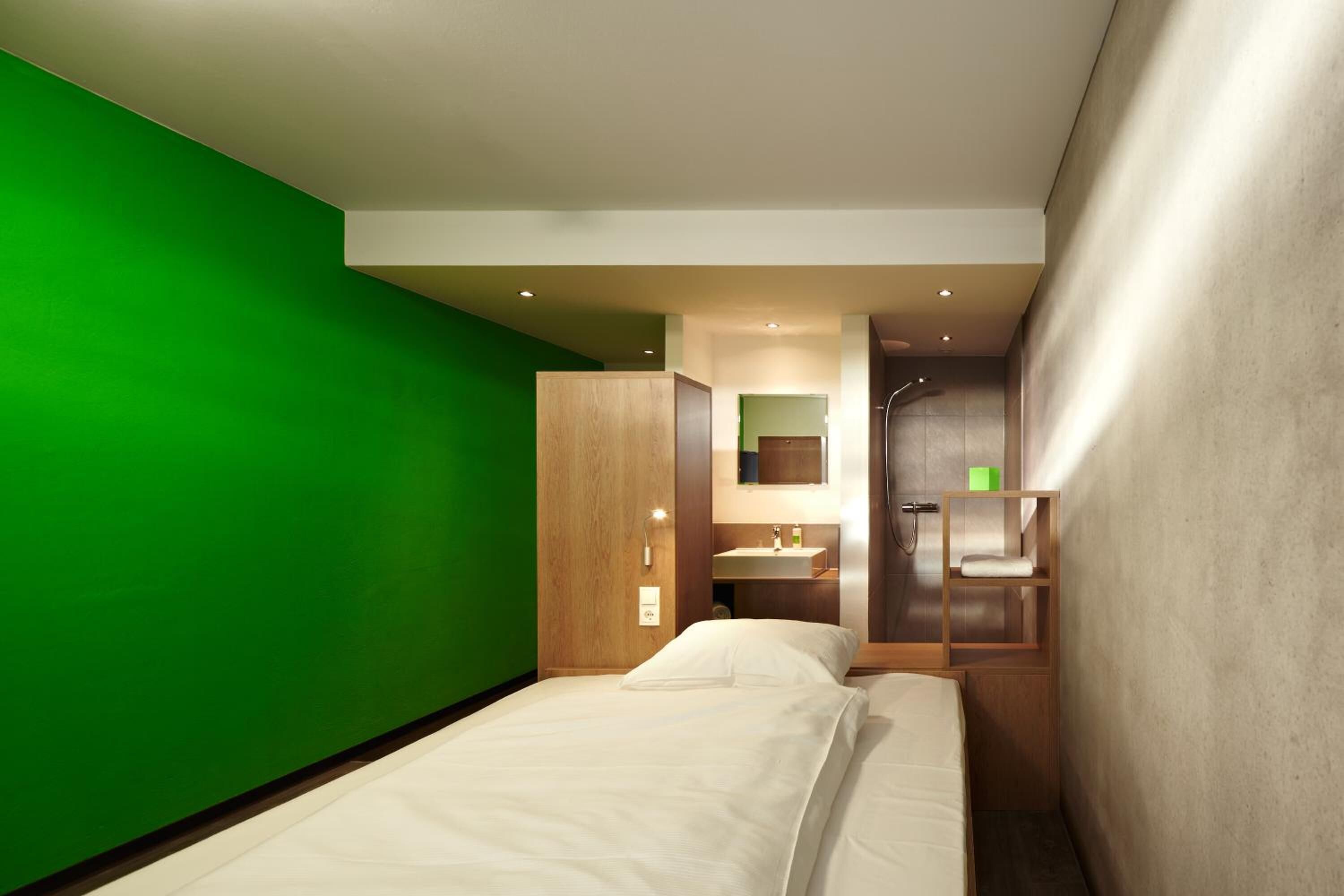 Una habitación de hotel moderna con una pared de acento verde, una cama con ropa de cama blanca, un armario de madera, y un baño con espejo, lavabo y ducha.