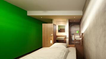 Una habitación de hotel moderna con una pared de acento verde, una cama con ropa de cama blanca, un armario de madera, y un baño con espejo, lavabo y ducha.