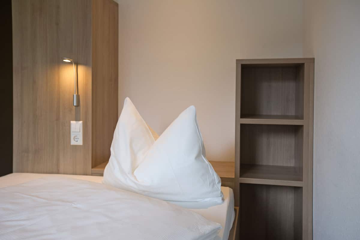 A neatly made bed with white linens and two pillows, a wall-mounted light, and a wooden shelf unit.