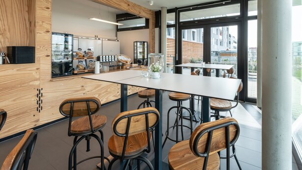 A modern café with wooden accents, a central white table, and bar stools. - a2 HOTELS DENKENDORF (Denkendorf)