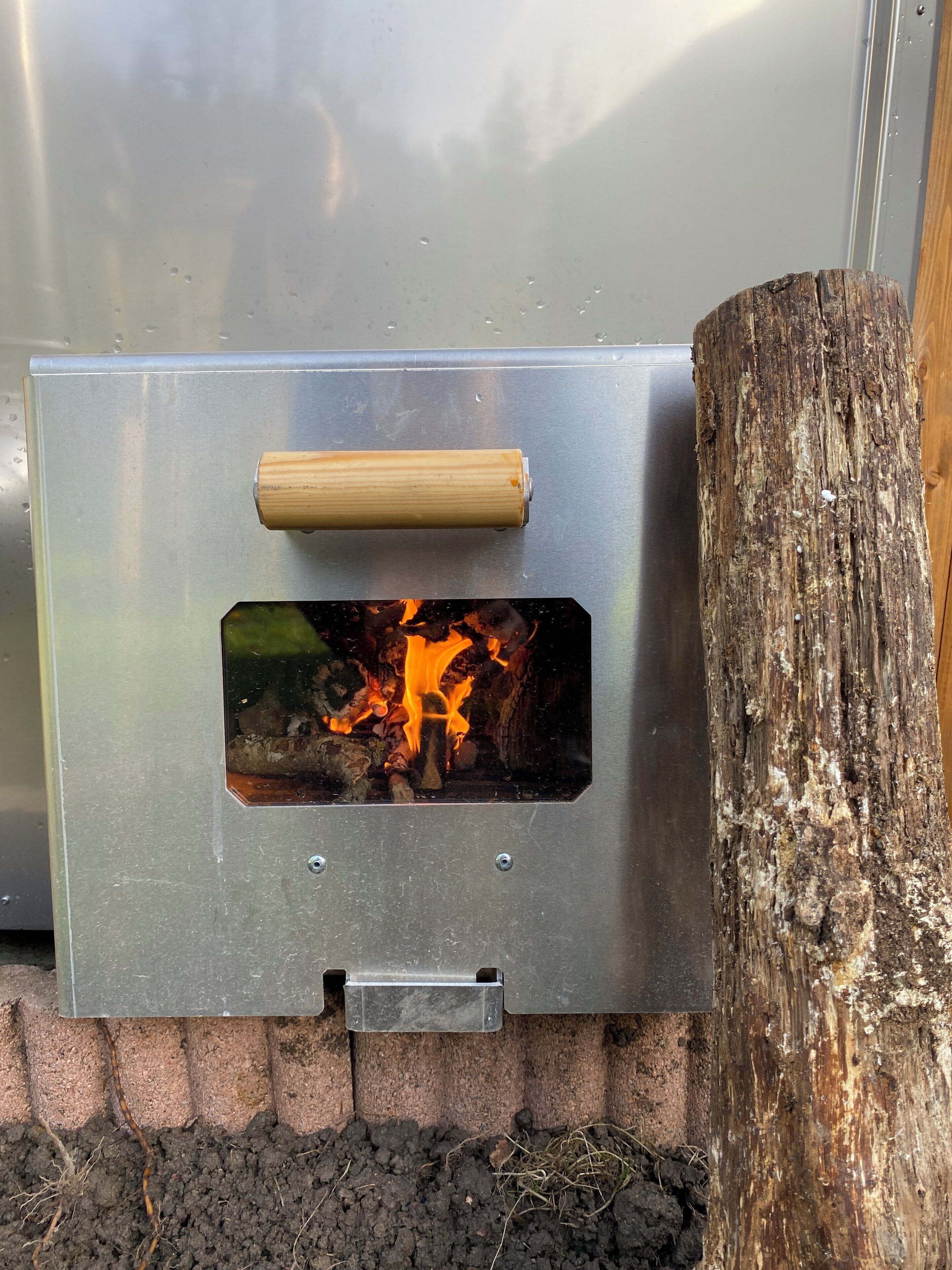 A metal grill with a wooden handle, a fire burning inside, and a tree stump beside it.