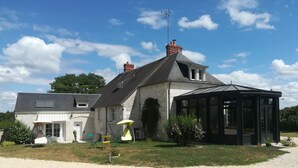 Une maison à deux étages dotée d’une grande véranda, d’une allée gravillonnée et d’un jardin aménagé avec du mobilier d’extérieur.