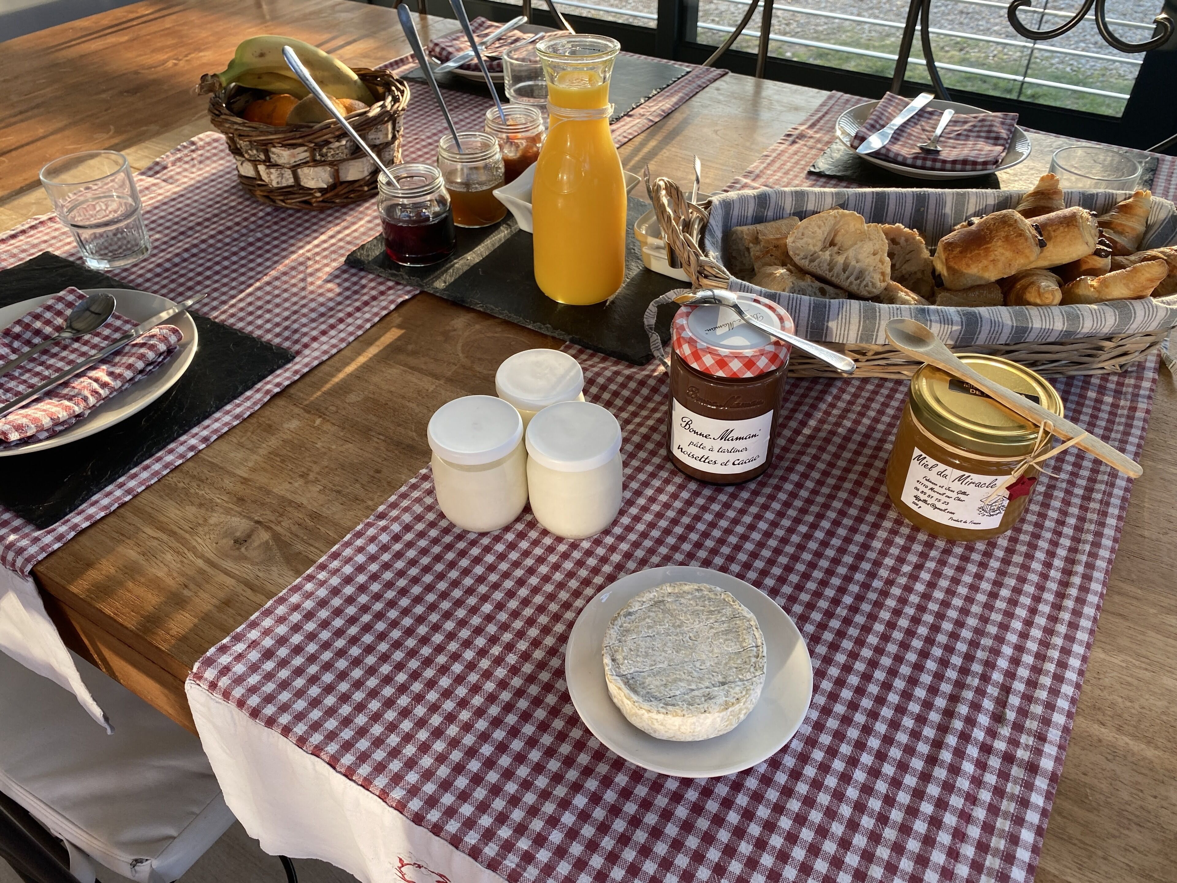 Ein Esstisch mit kariertem Tischtuch, ein Korb mit Croissants, eine Honigdose und ein Glas Orangensaft.