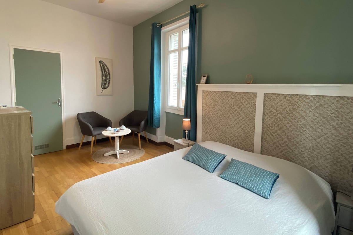 Ein Schlafzimmer mit einem Bett, zwei Stühlen, einem kleinen Tisch, einem Deckenventilator, einem Fenster mit Vorhängen und einer Tür.