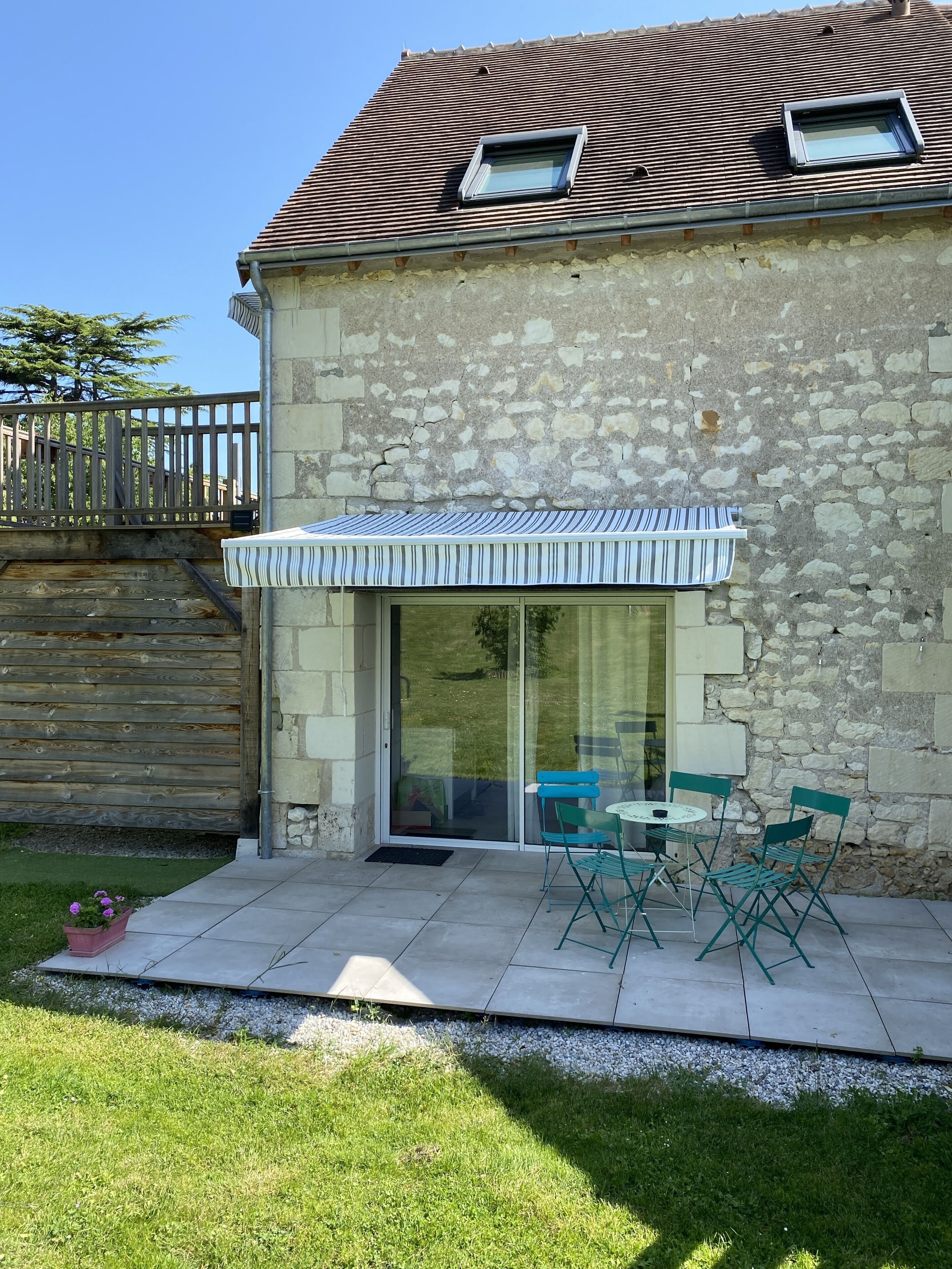 A stone house with a tiled roof, a covered patio with a table and chairs, and a wooden fence in the background.