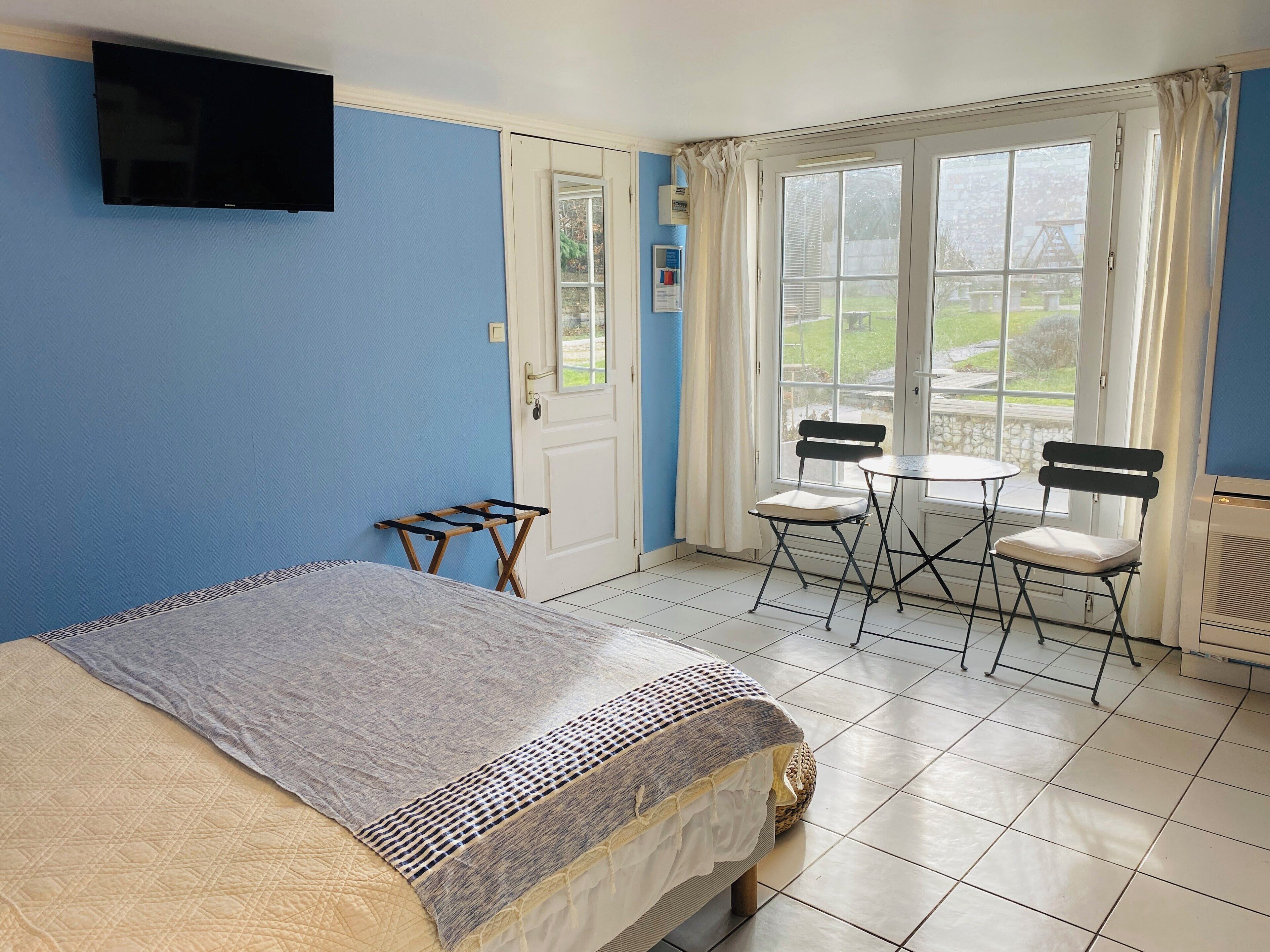A room with a bed, a small table with chairs, a TV mounted on the wall, and a window with curtains.