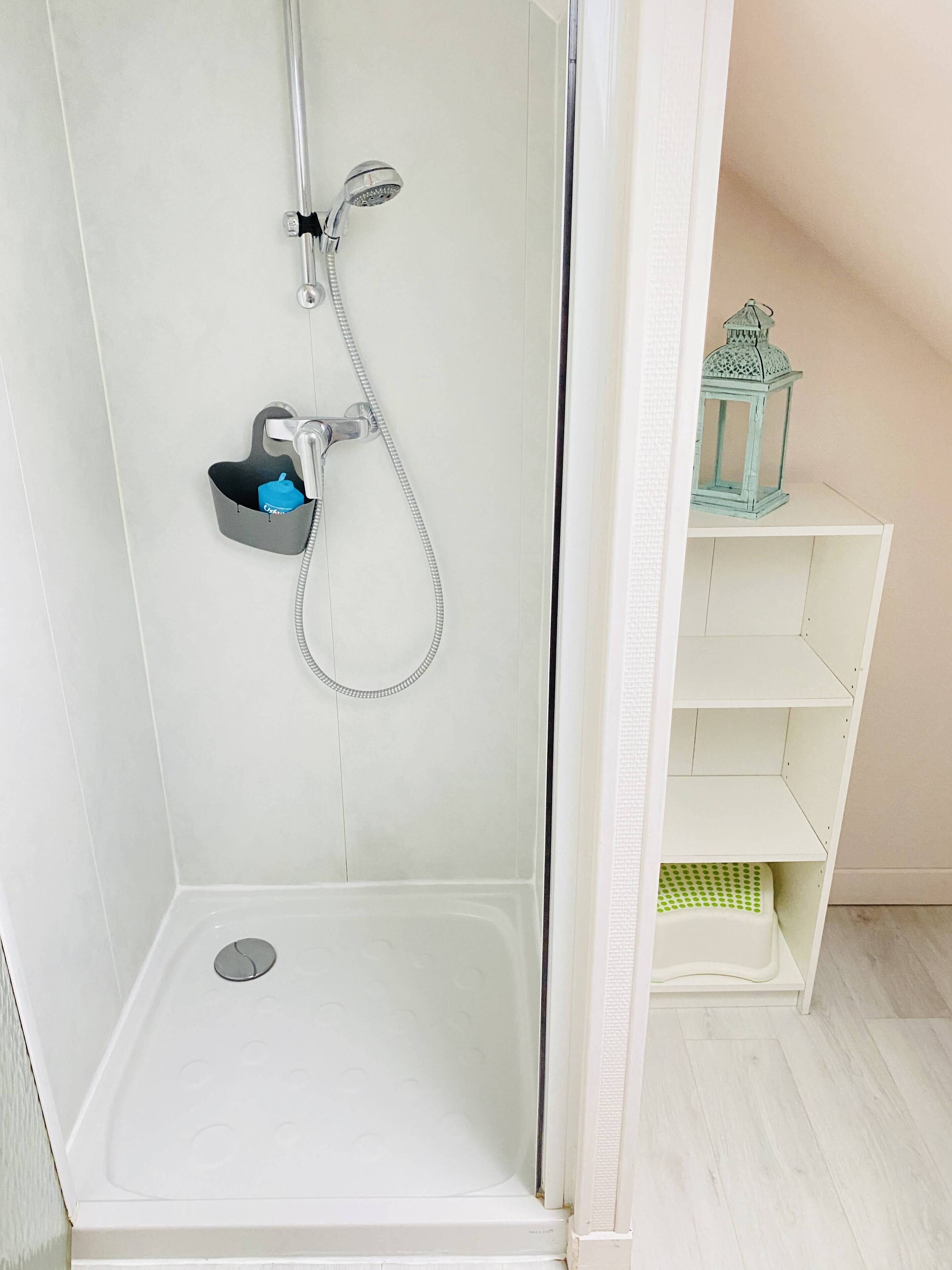 Une douche équipée d’un pommeau de douche à main et d’un robinet mural, une petite étagère avec une bouteille bleue et un mur recouvert de carreaux blancs.