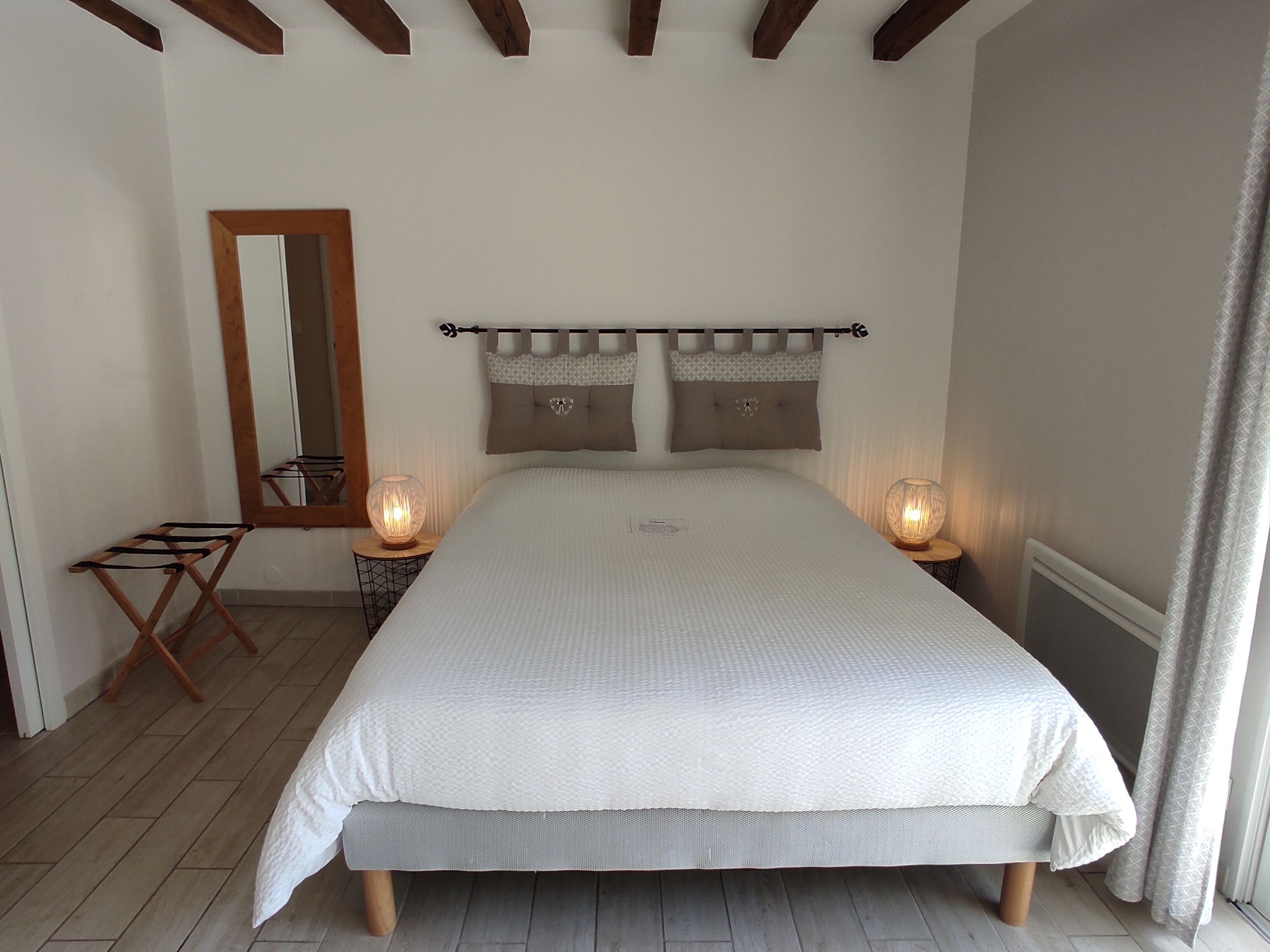 A bedroom with a large bed, two bedside lamps, a wooden chair, a mirror, and a window with curtains.