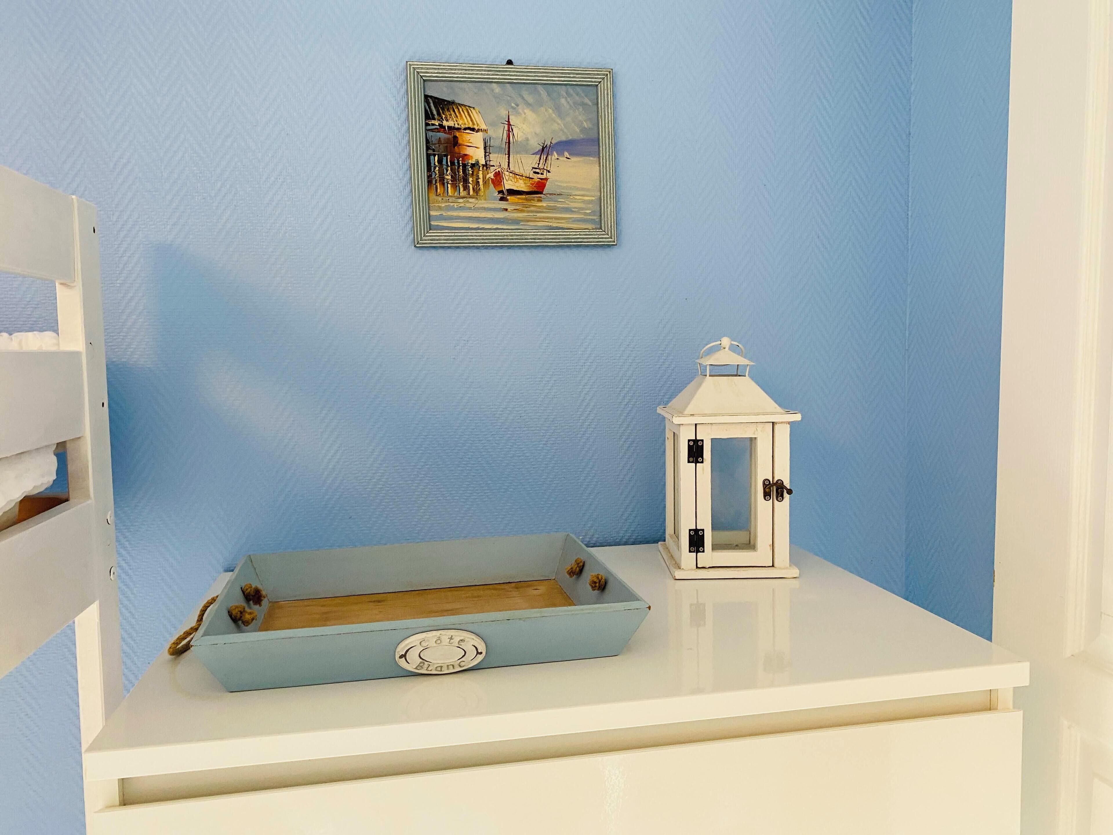 A white dresser with a blue tray, a white lantern, and a framed painting on a blue wall.