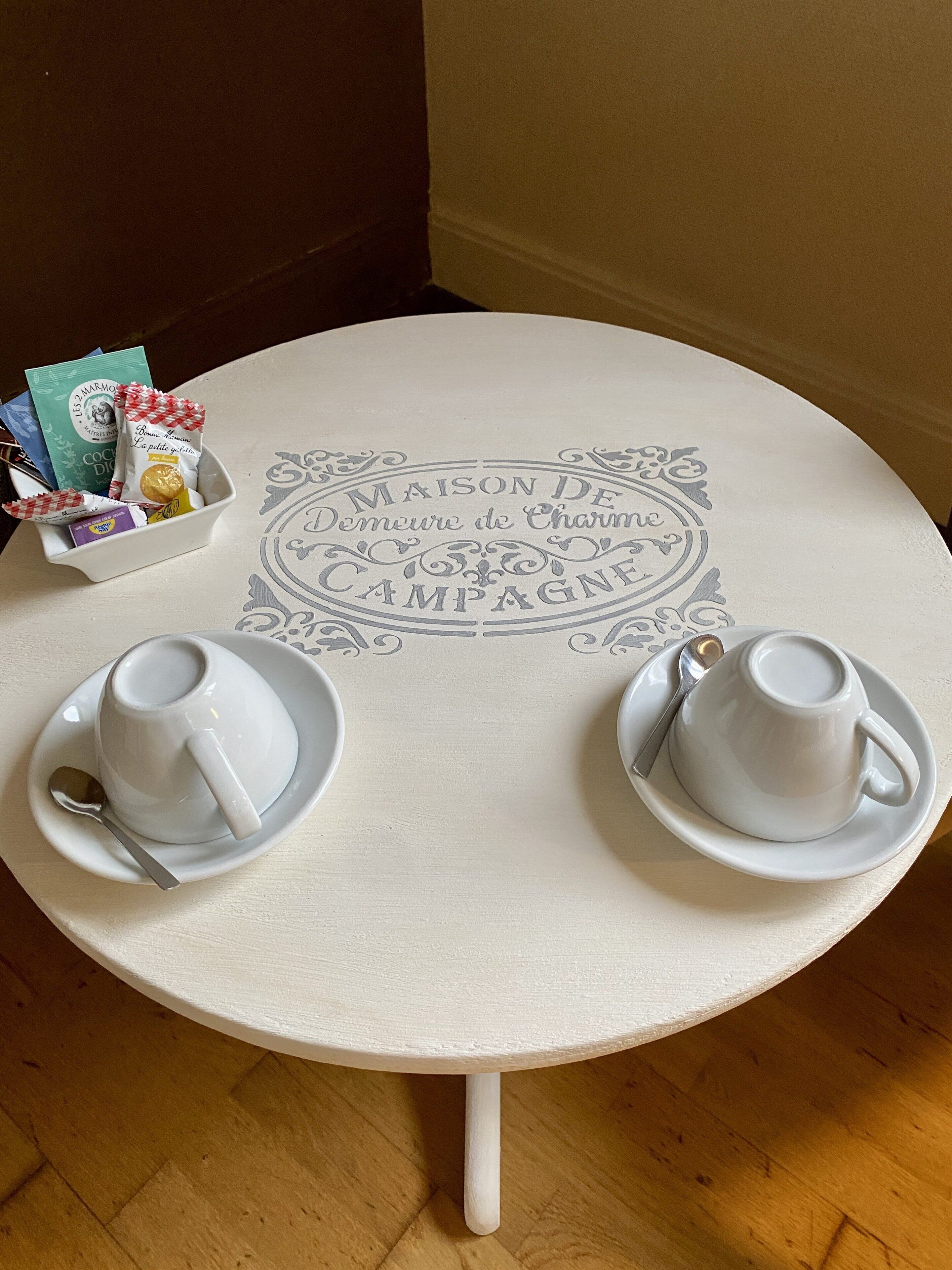 A small round table with two white cups and saucers, a spoon, and a bowl containing various packaged snacks.