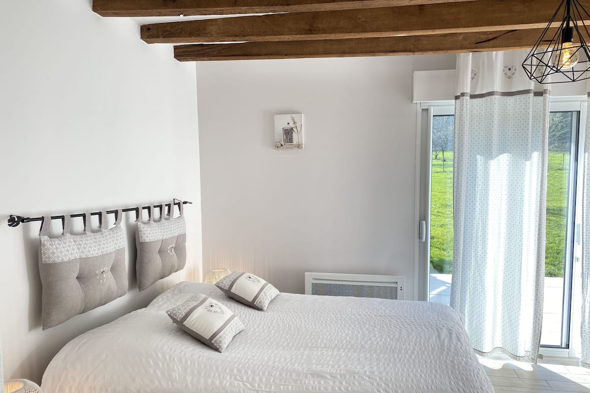 Ein Schlafzimmer mit einem großen Bett, einer Kopfteil mit vielen Kissen, einer Nachttischlampe und Blick ins Grüne.