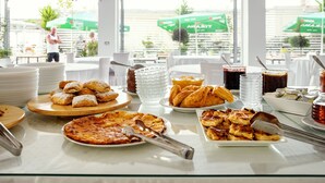 Ein Frühstücksbuffet mit verschiedenen Backwaren, einer Pizza und Getränken auf einem Tisch in einem hellen Raum mit großen Fenstern.