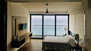 Ein Schlafzimmer mit großem Fenster und Meerblick, einem Bett, einem Nachttisch und einem an der Wand befestigten Fernseher.