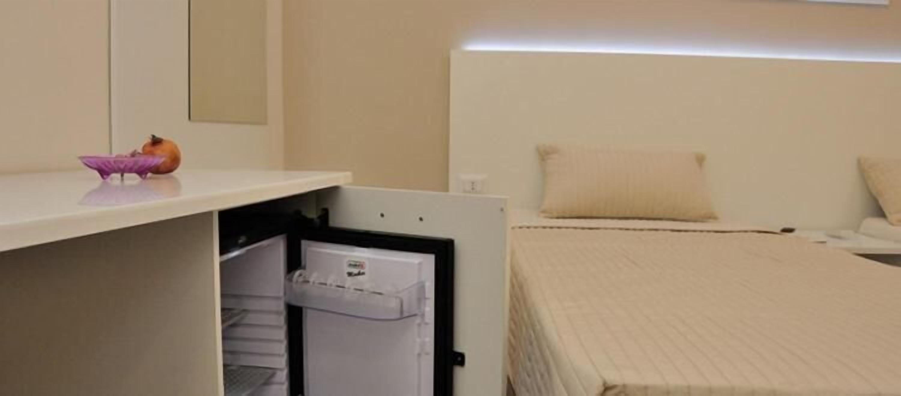 A hotel room with a bed, a bedside table, a small refrigerator, and a wall-mounted shelf with a decorative item.