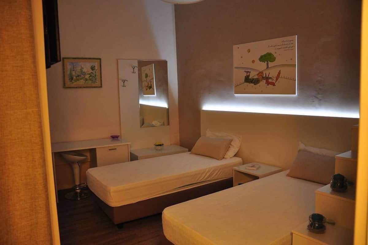 A hotel room with two single beds, a desk, a chair, a mirror, and artwork on the walls.