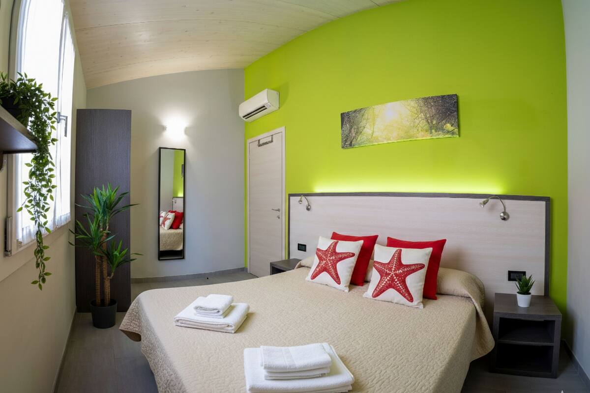 A neatly made bed with white and red pillows, a bedside table with a cup, and a small potted plant.