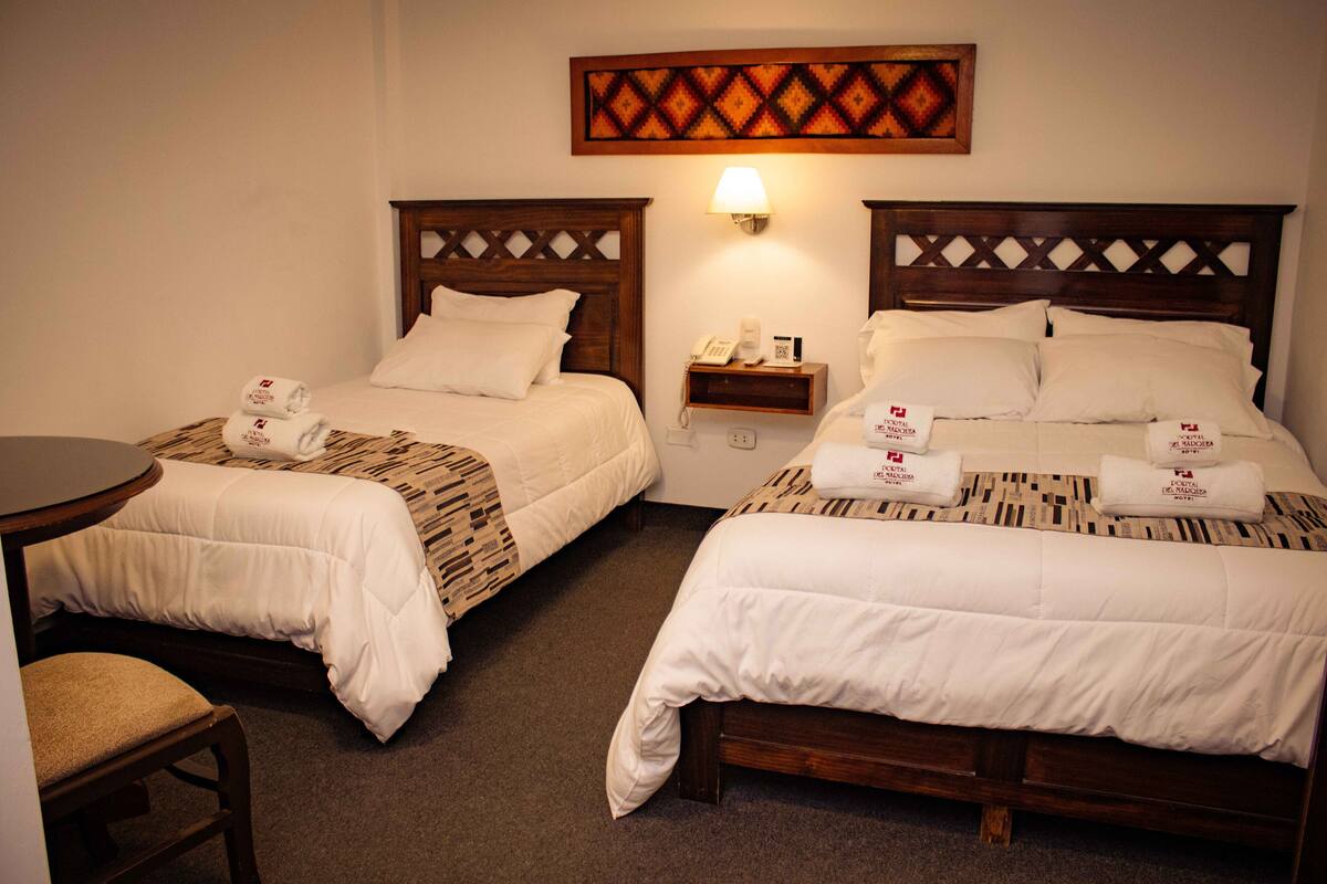 A hotel room with two beds, each with a wooden headboard, a bedside table, and a chair. There is a wall-mounted lamp and a small shelf with a phone and a book.